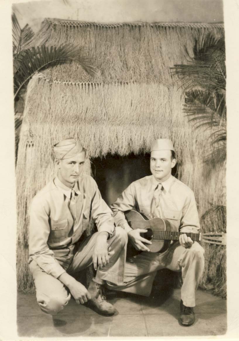 2 men, one with guitar, squatting in front of straw hut photograph