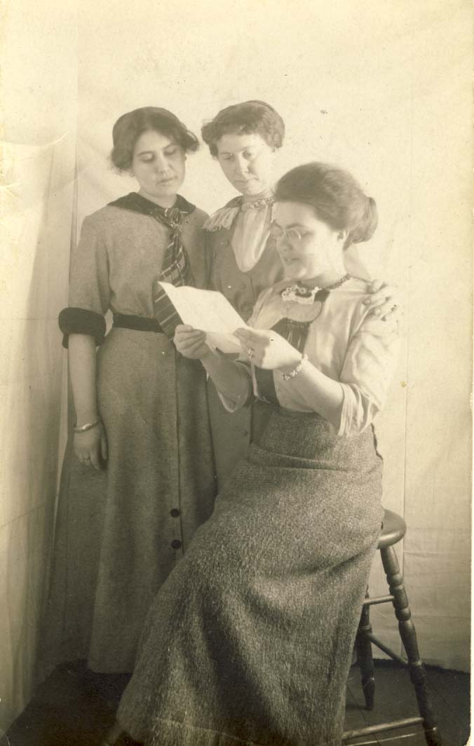 Three women, one seated on stool while reading a letter postcard